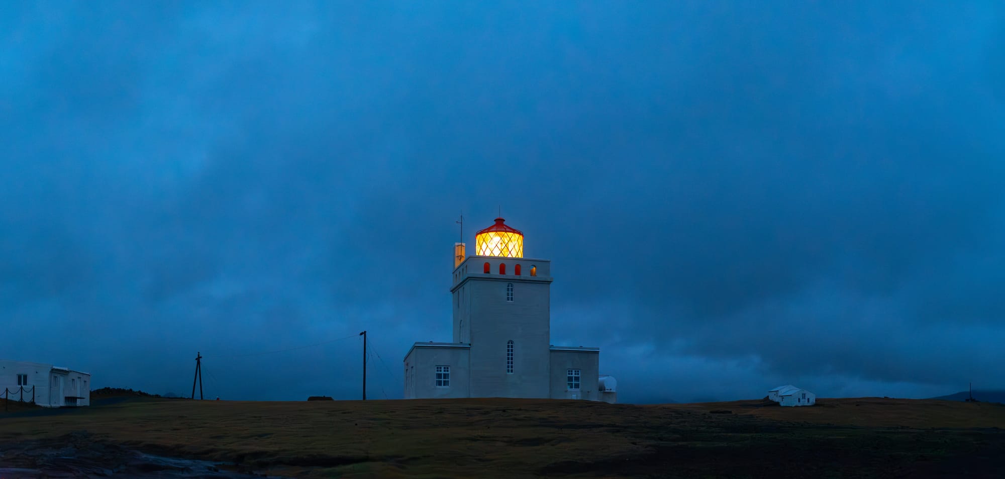 A brightly lit lighthouse standing against a dark, moody sky at dusk or dawn, with its warm light contrasting sharply with the deep blue tones of the surrounding landscape and distant buildings.