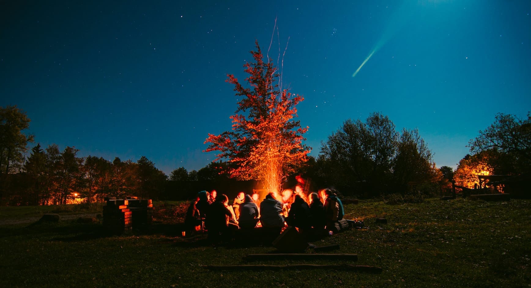 A group of people gathers around a large bonfire at night under a starry sky with a comet visible in the distance taken by European Elopement Photographer Sturmsucht.