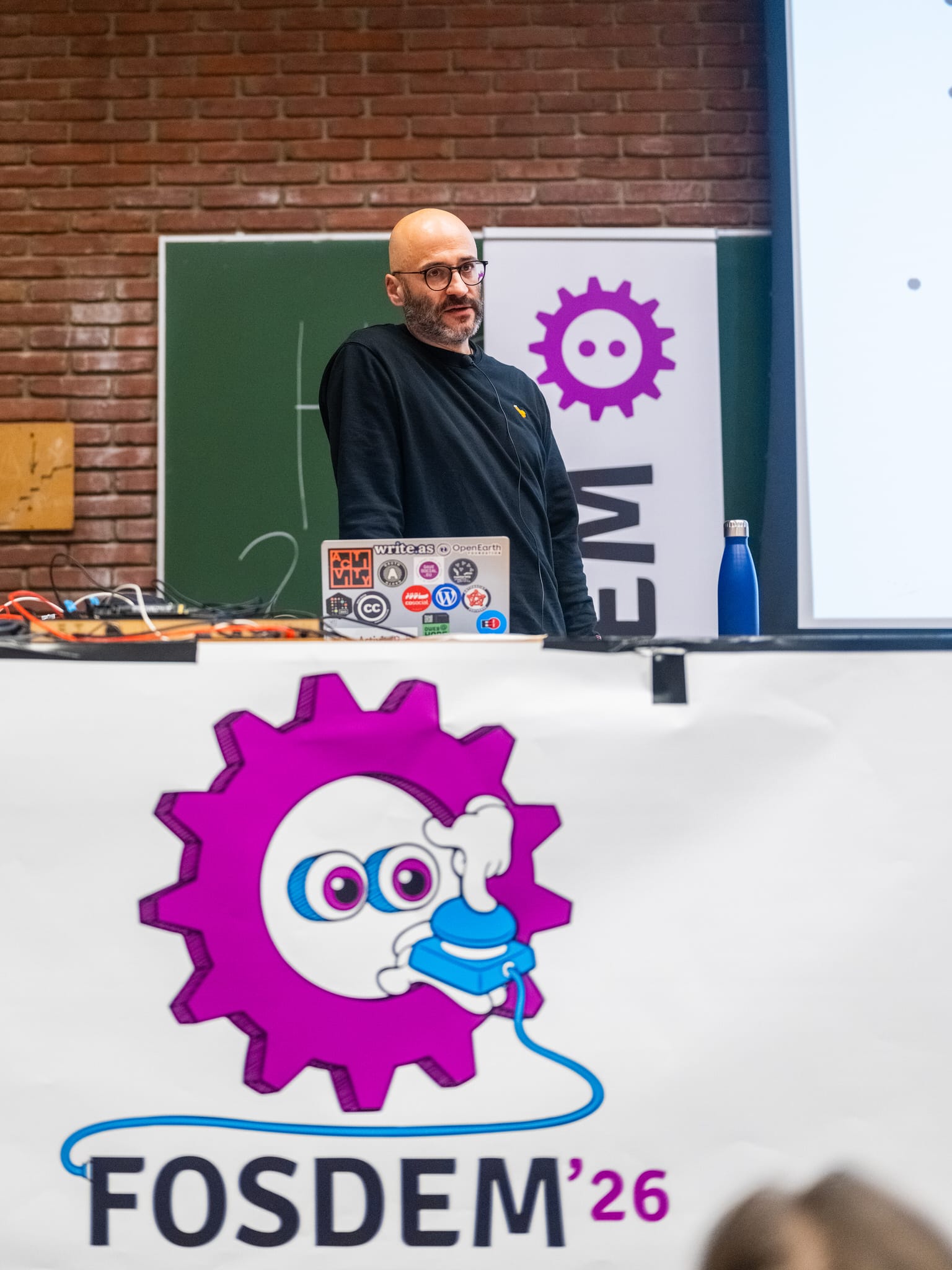Matthias with a beard and glasses stands behind a laptop with stickers, while presenting in front of a brick wall, a blackboard, and a "FOSDEM'26" banner with a purple gear logo.