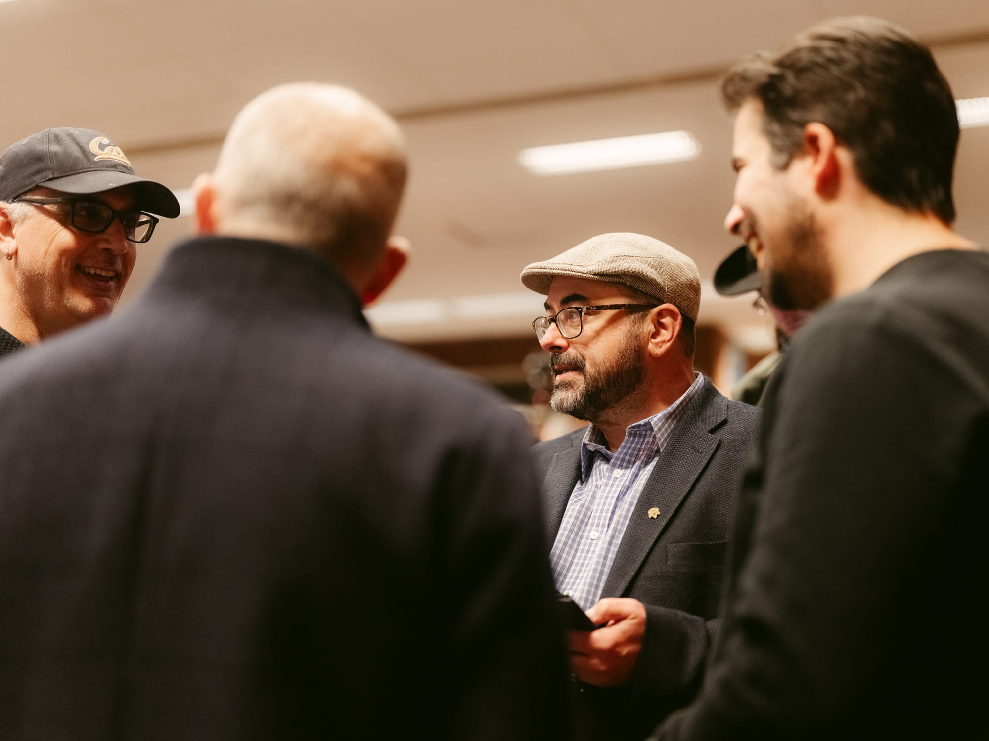 Four men, two wearing hats and glasses, are engaged in a lively conversation indoors, with one smiling broadly on the left and another with a beard and flat cap in the center.