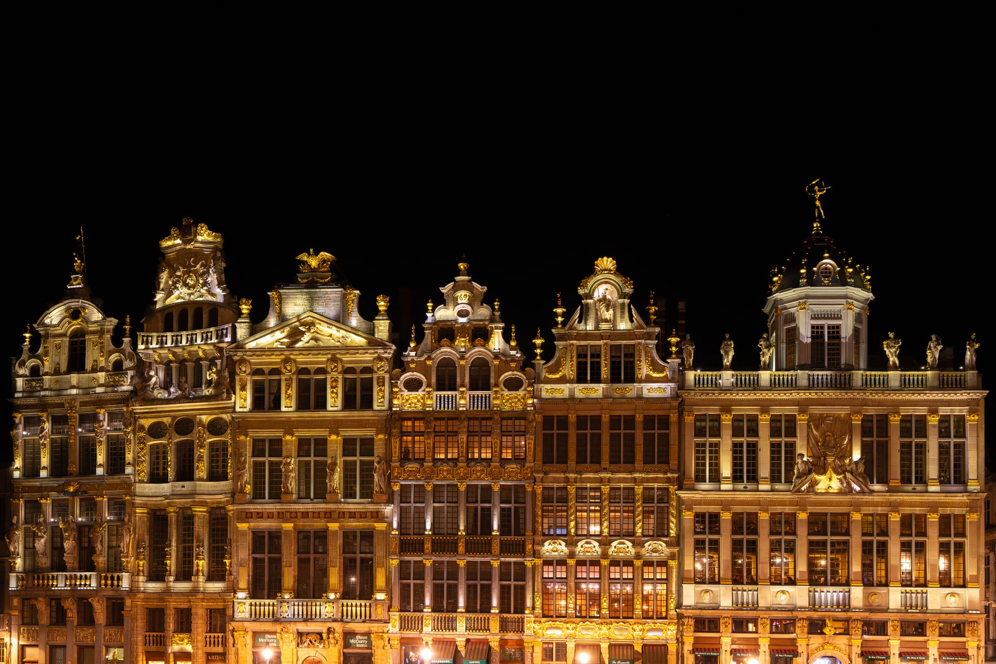 The image captures a row of grand, ornate buildings in Brussels, illuminated at night with warm golden lights against a dark sky, showcasing their intricate architectural details and statues.