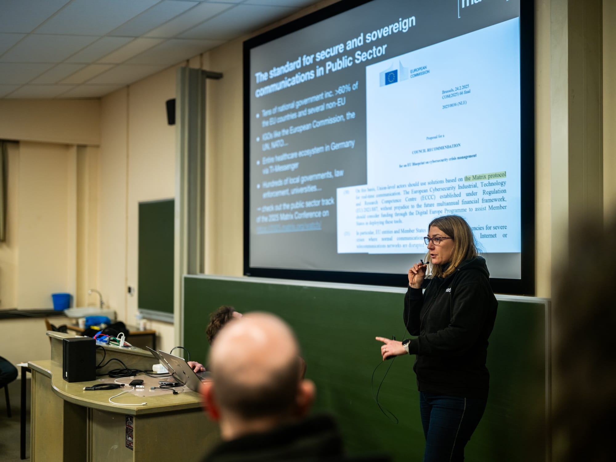 Amandine from Matrix speaks into a microphone while presenting in a lecture hall, with a slide titled "The standard for secure and sovereign communications in Public Sector" visible on a large screen behind her.