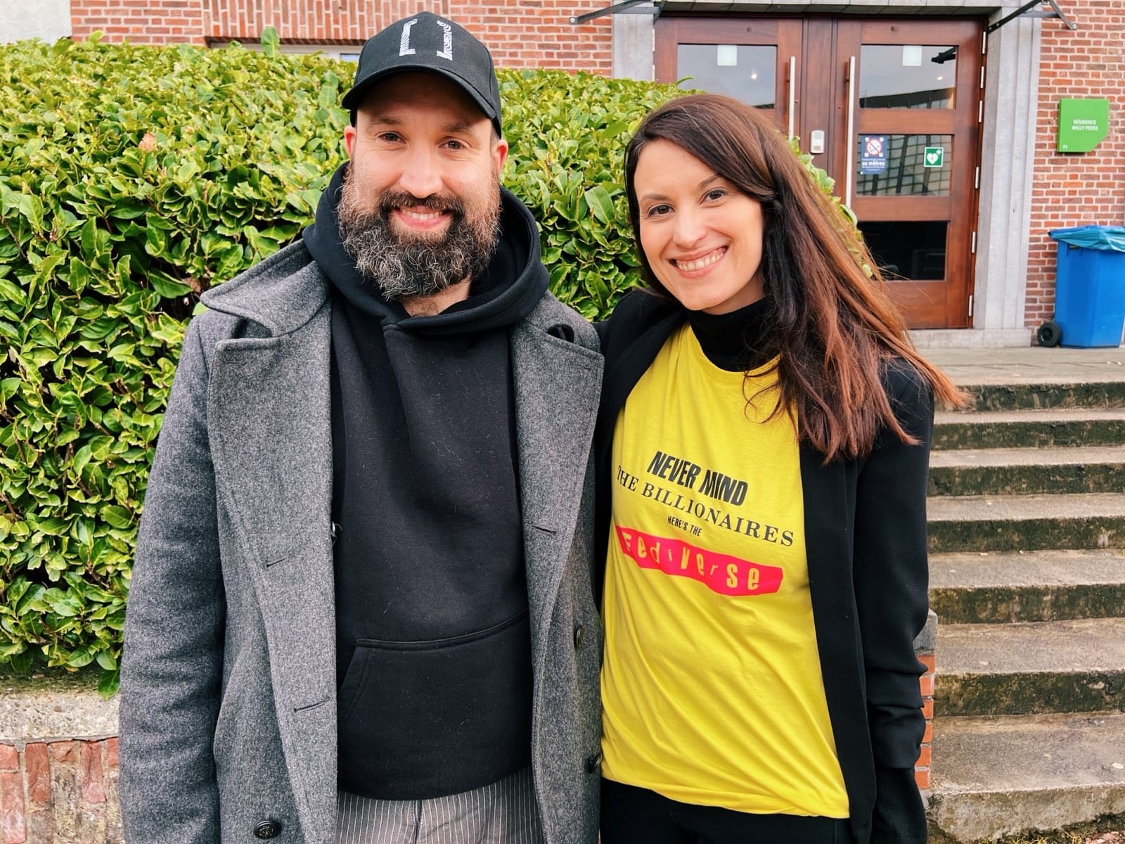 A smiling bearded man in a cap and grey coat stands next to a smiling woman in a yellow "Never Mind the Billionaires Here's the Fediverse" t-shirt, both posing outdoors in front of a green bush and a building.