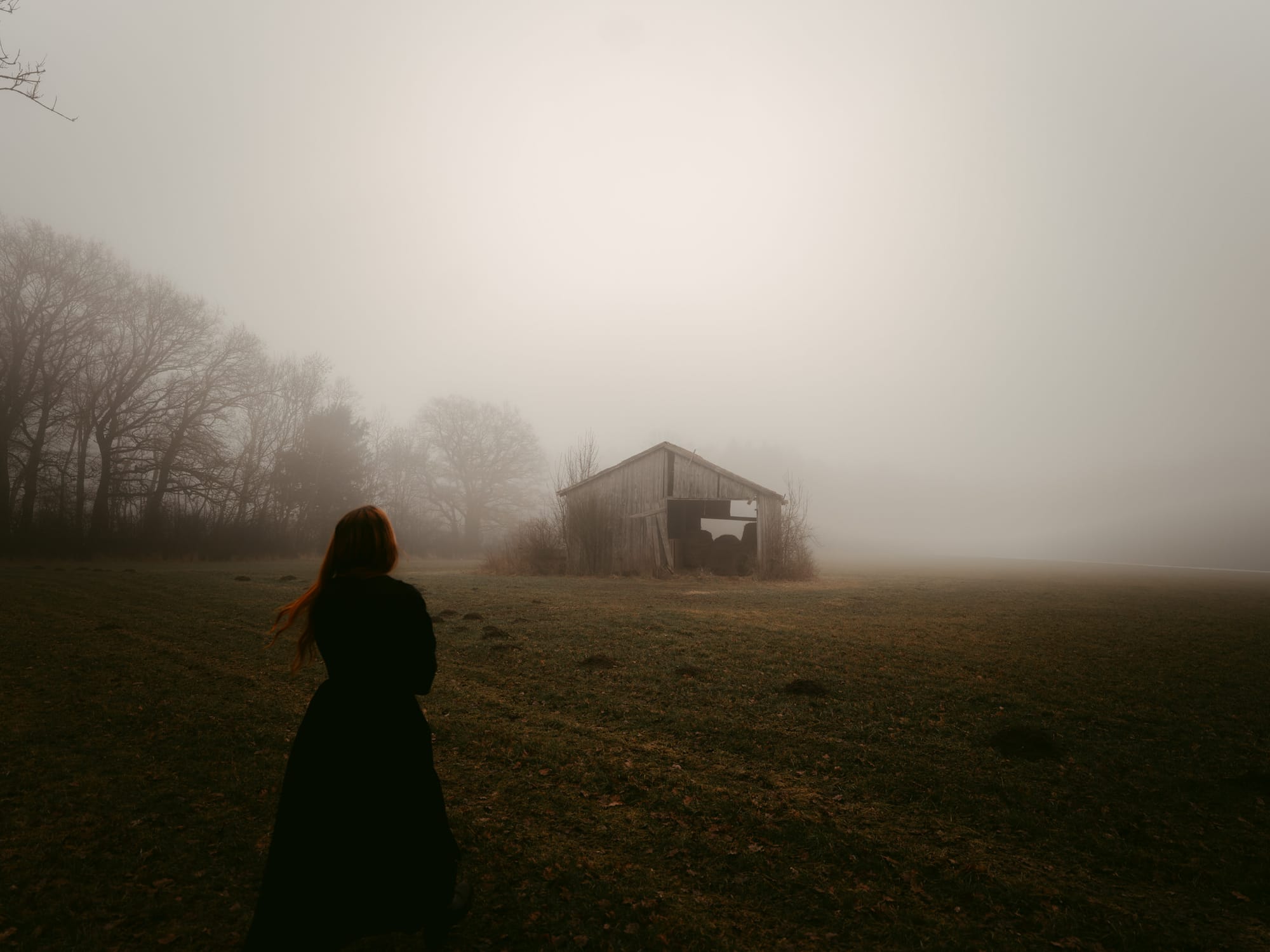 An image taken by European Elopement Photographer Sturmsucht shows a young women with red hair, seen from behind, stands in a foggy field looking towards an old, dilapidated barn. Bare trees are visible in the mist to the left.