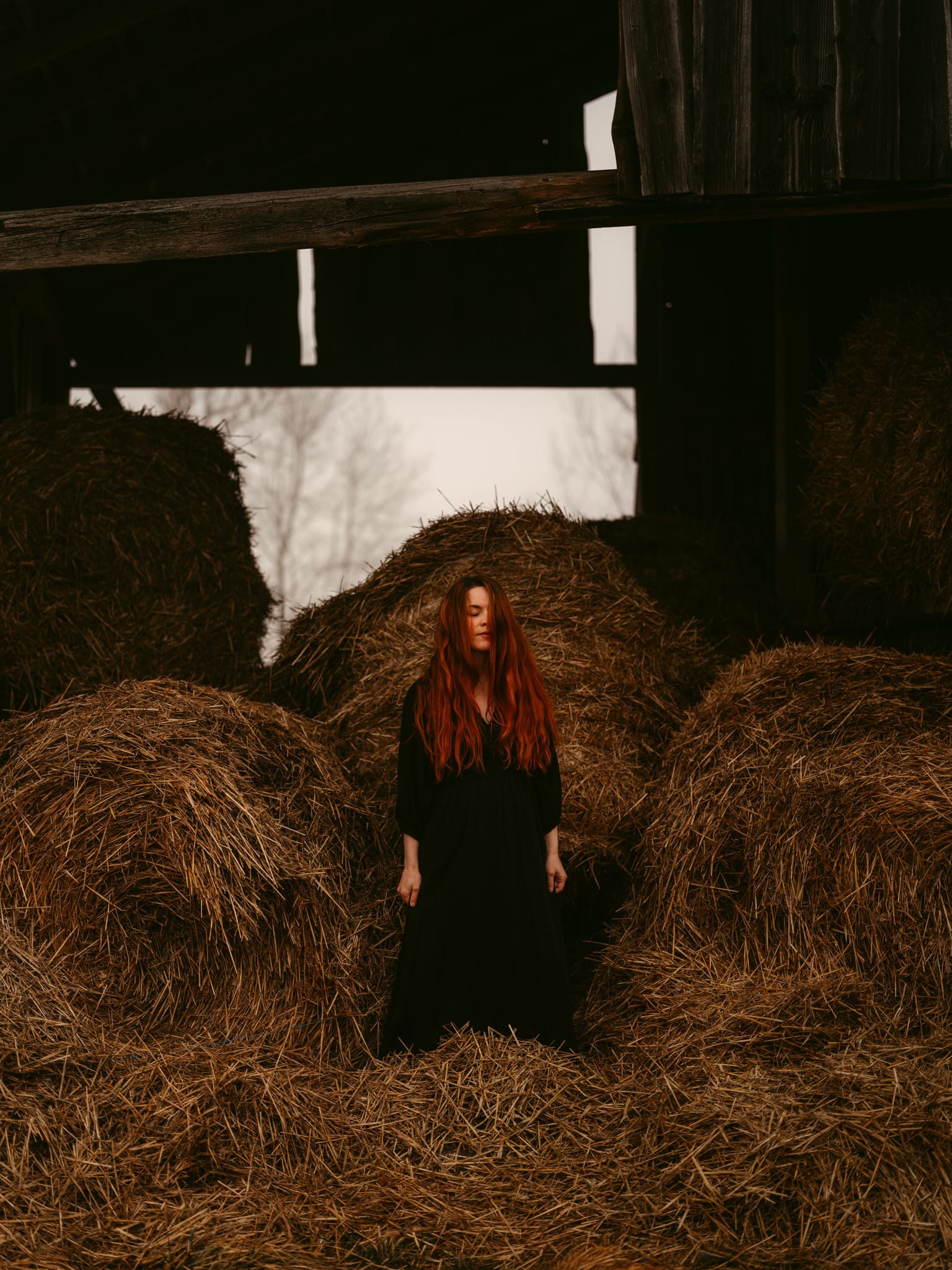 A young woman with long red hair and a dark dress stands with closed eyes amidst large hay bales inside a barn, caputred by European Elopement Photographer Sturmsucht