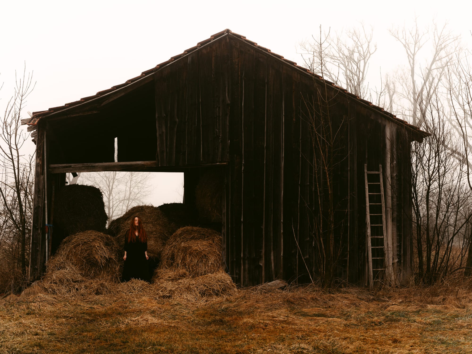 European Elopement Photographer Sturmsucht captured a young woman with long red hair and a dark dress sits on hay bales inside a weathered wooden barn, which stands in a foggy, muted landscape with bare trees.