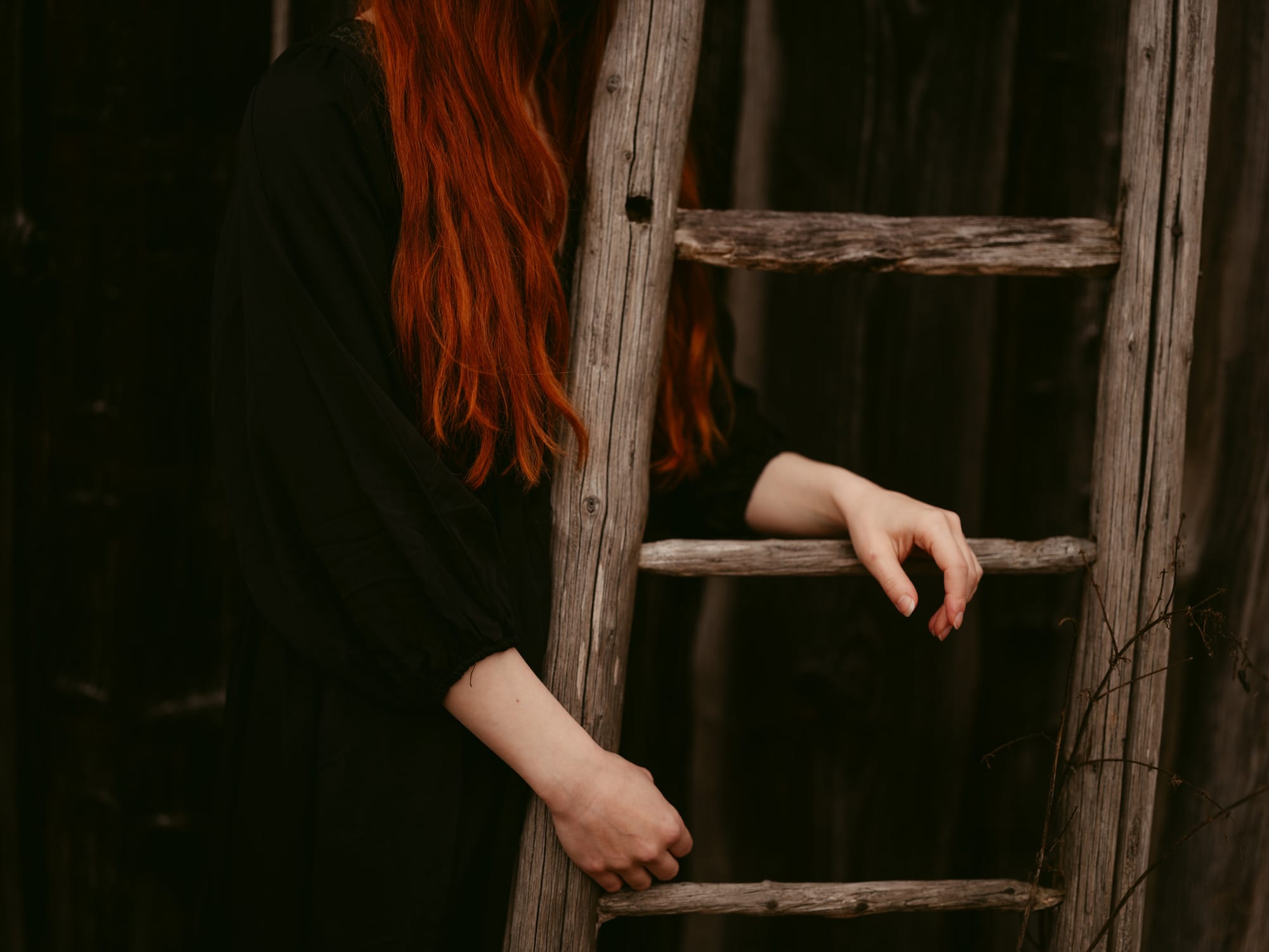 A close-up of a young woman with long red hair and a dark dress, with their hands resting on an old wooden ladder during a session with European Elopement Photographer Sturmsucht
