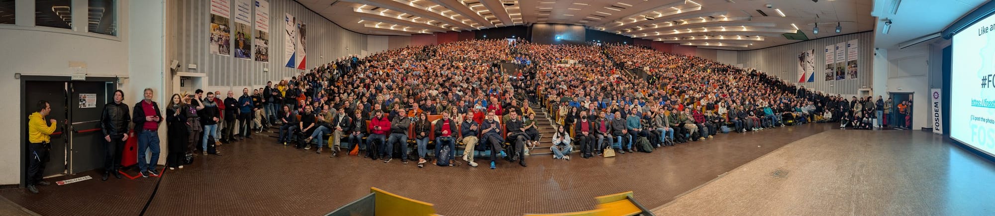 This image shows a panoramic view from a speaker's perspective at FOSDEM, capturing a large, brightly lit auditorium filled with a diverse, casually dressed audience seated in tiered rows, with banners, a visible "FOSDEM" projection screen on the right, and an open stage area, conveying the scale and energy of the event.