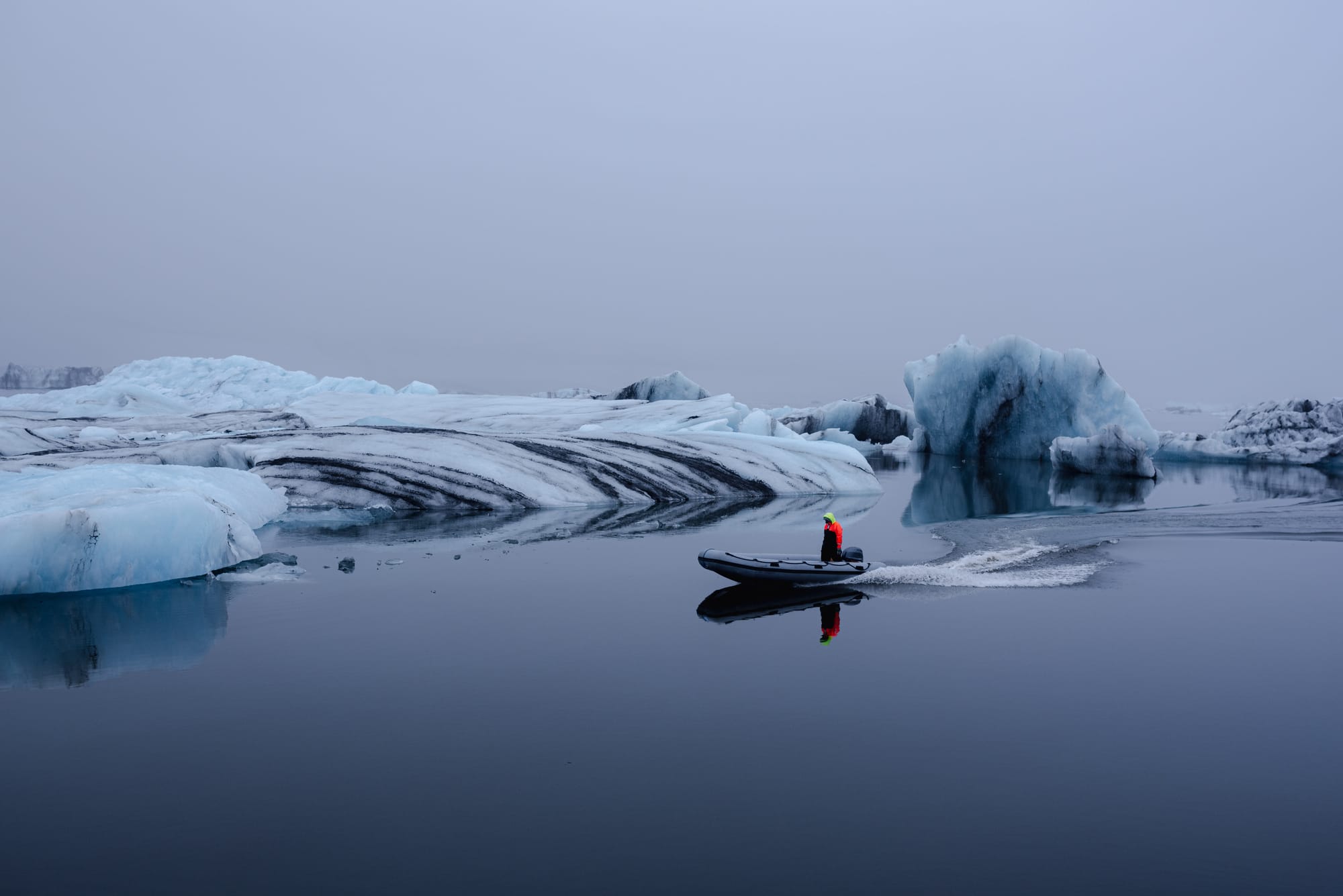 A person in a red jacket with a neon yellow hood, riding a small boat across an Icelandic glacier lake.