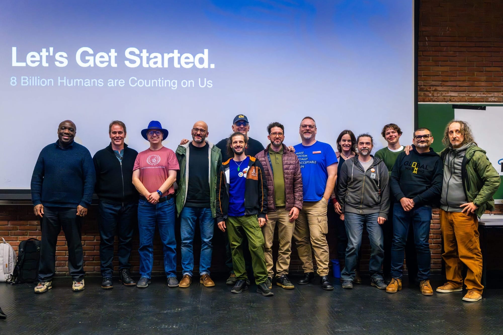 This image by Chris Zielecki / Sturmsucht captures a group of 13 speakers on a stage at FOSDEM with a large screen displaying: Let's Get Started. 8 Billion Humans are Counting on Us